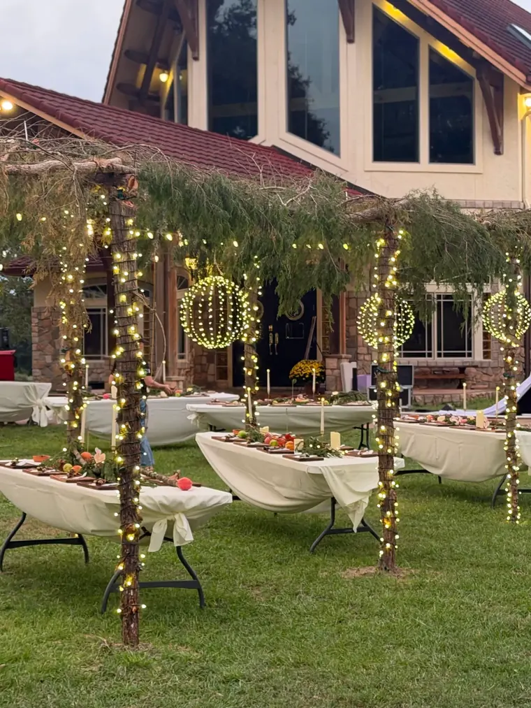 Lit evergreen garland arch over a lawn in daylight, with wrapped tree trunks and banqueting tables.