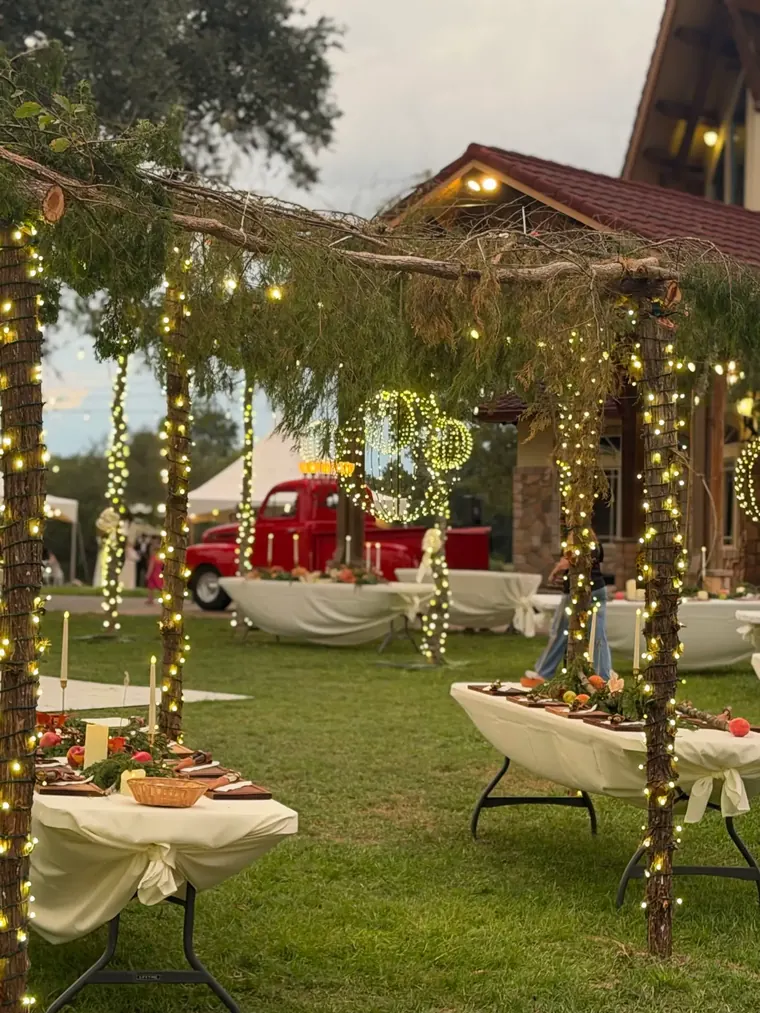 Wide view of a lawn with lit arches, wrapped trees and tables during setup.