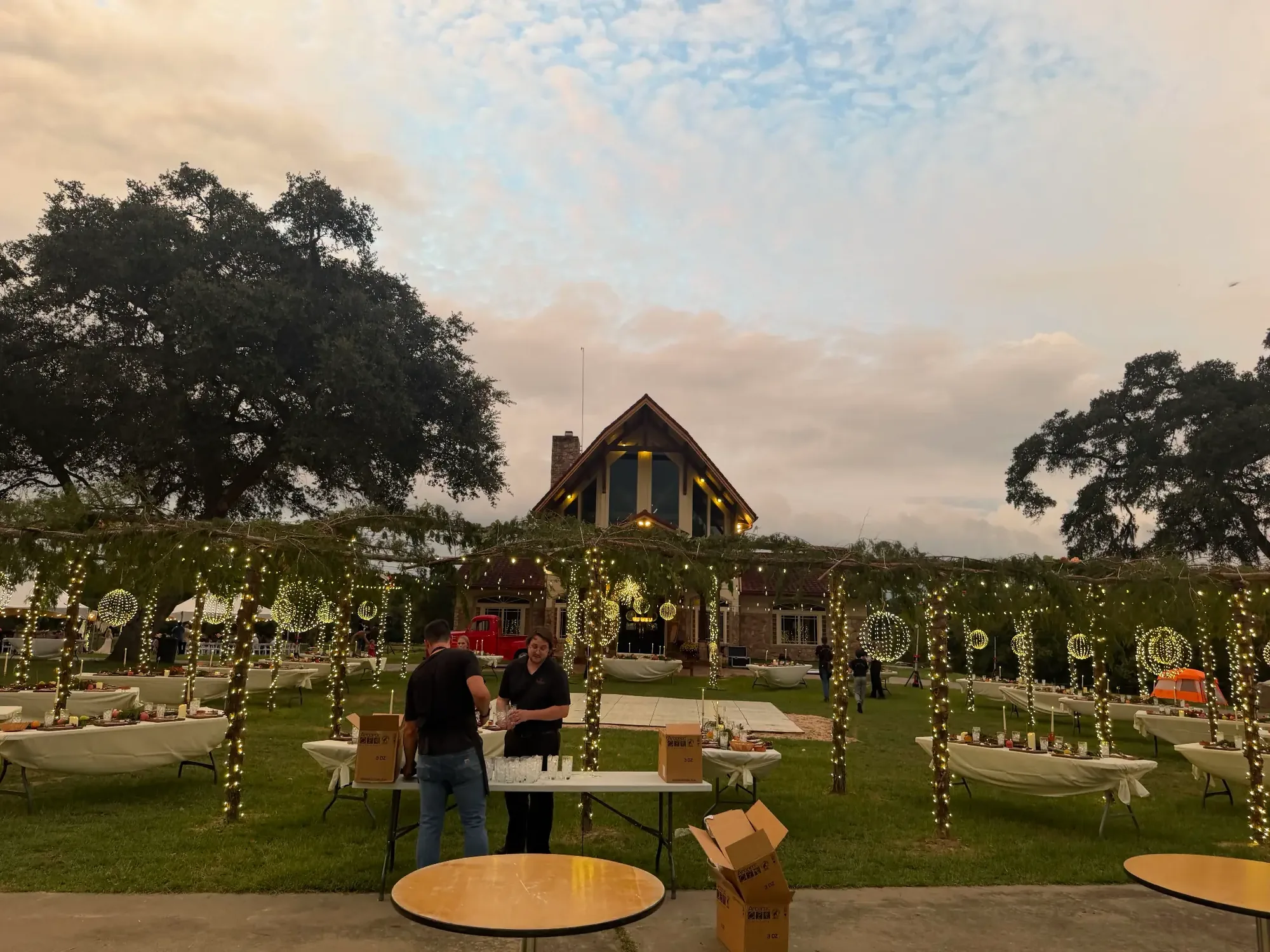 Event lawn at dusk with a string-light canopy strung between mature trees.