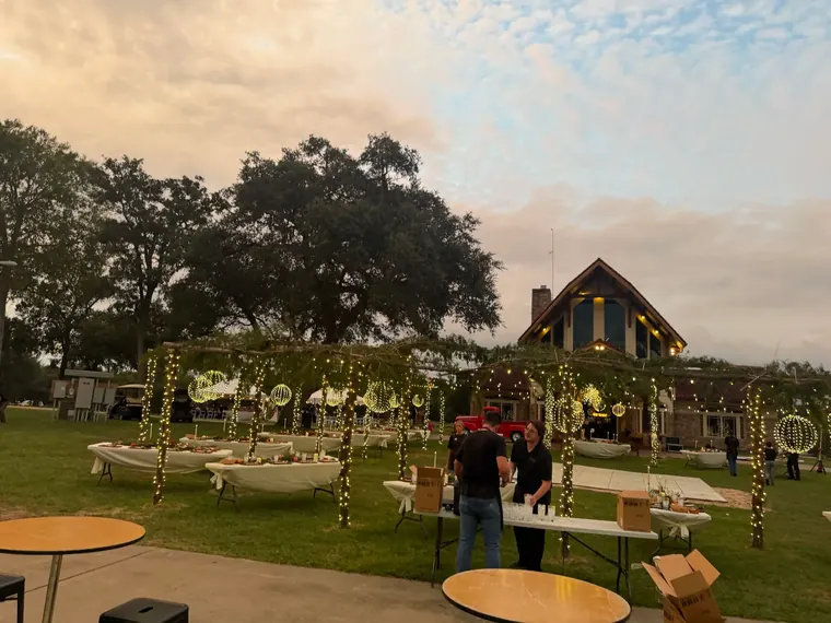 Event lawn at dusk with wrapped trees, lit arches and guests arriving.