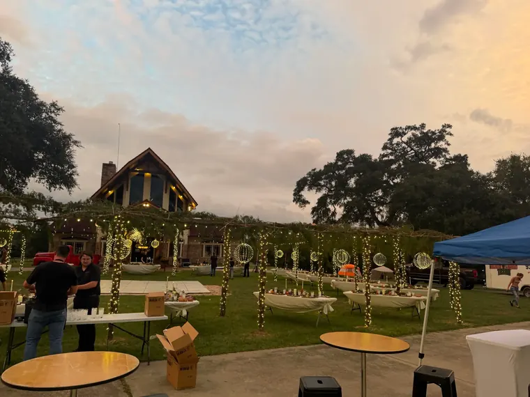 Wide dusk view across an event lawn with lit trees, tables and a timber building.