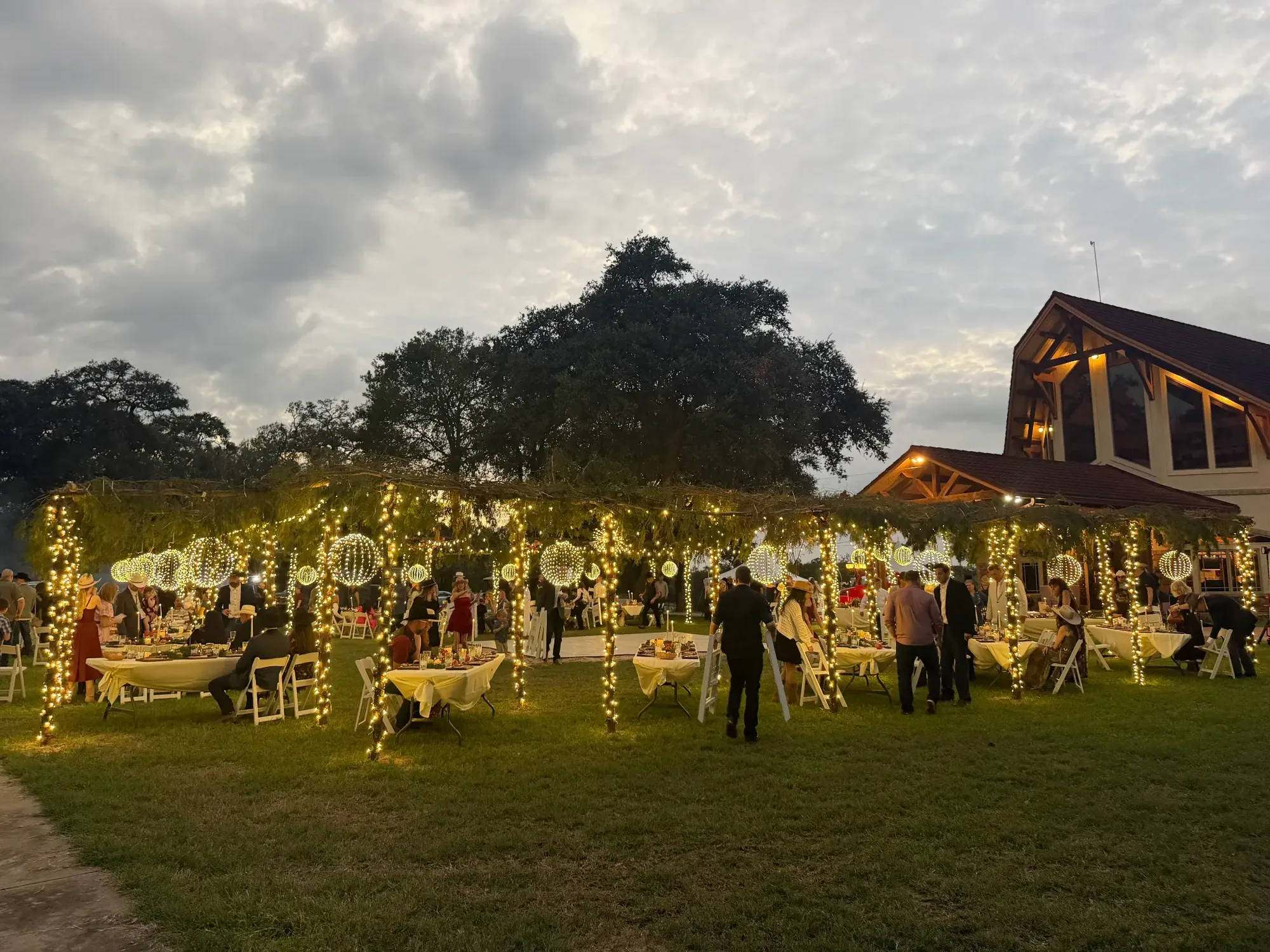 Night view across a crowded event lawn with wrapped trees and lit garland arches.