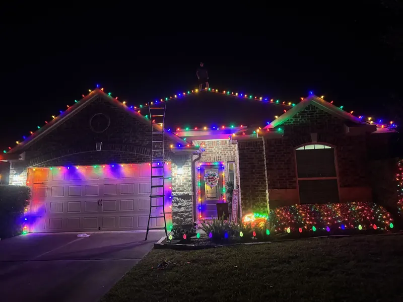 Brick home at night with multicolor C9 bulbs along the gables, garage door and front door, a wreath and lit shrubs.