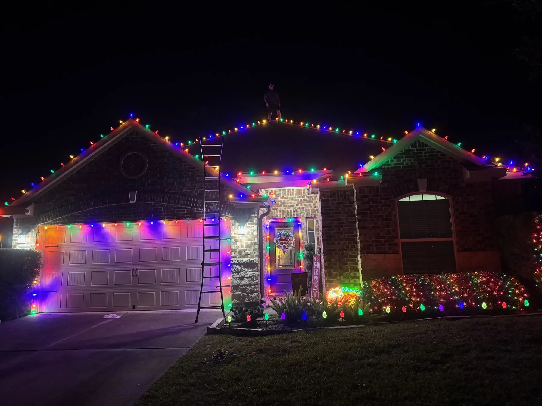 Brick home at night with multicolor C9 bulbs along the gables, garage door and front door, a wreath and lit shrubs. Representative of installs like those near Katy.