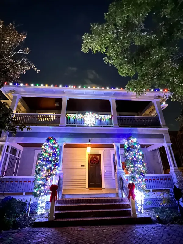 Two-story craftsman home with multicolor lights on the roofline, lit garland and a wreath on the balcony, and two glowing evergreen topiaries flanking the porch steps.