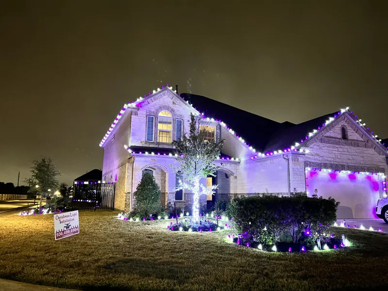 Two-story limestone home at night with purple and white C9 lights on the gables and garage, a white-wrapped tree and purple and white lights around the beds.