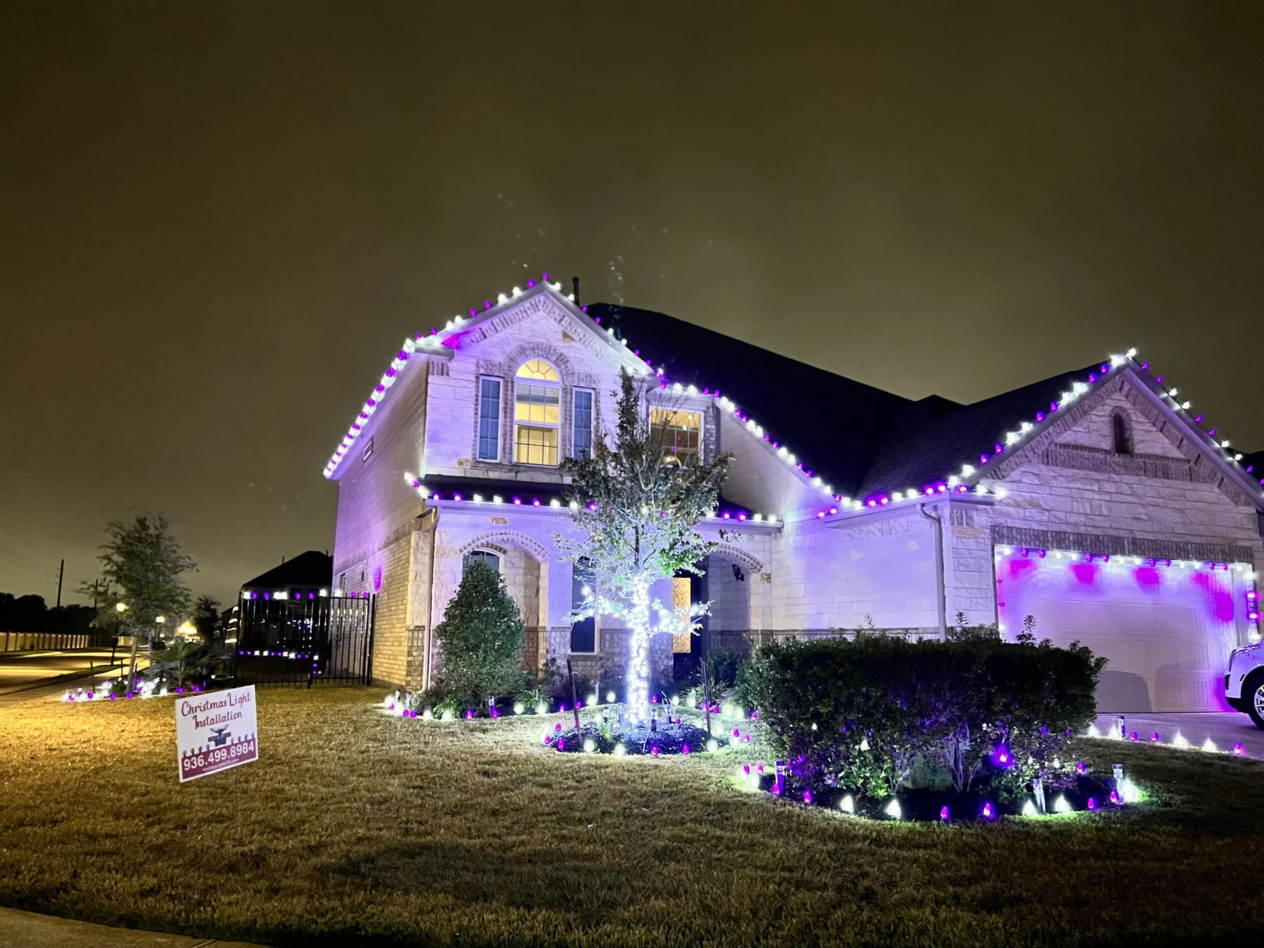 Two-story limestone home at night with purple and white C9 lights on the gables and garage and a white-wrapped tree in the front bed.