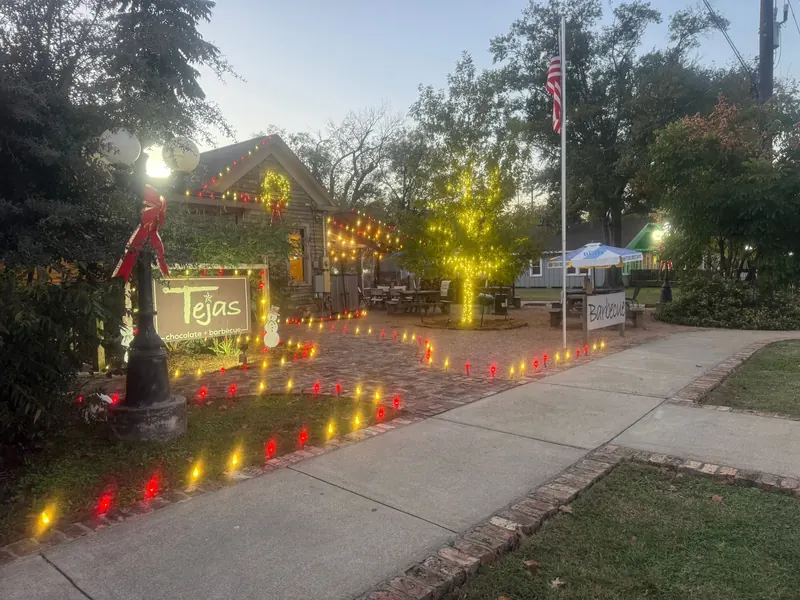 Cafe courtyard at dusk with red and amber C9 lights tracing a brick pathway, a glowing wrapped tree, a lit wreath and bows on the lamp post.