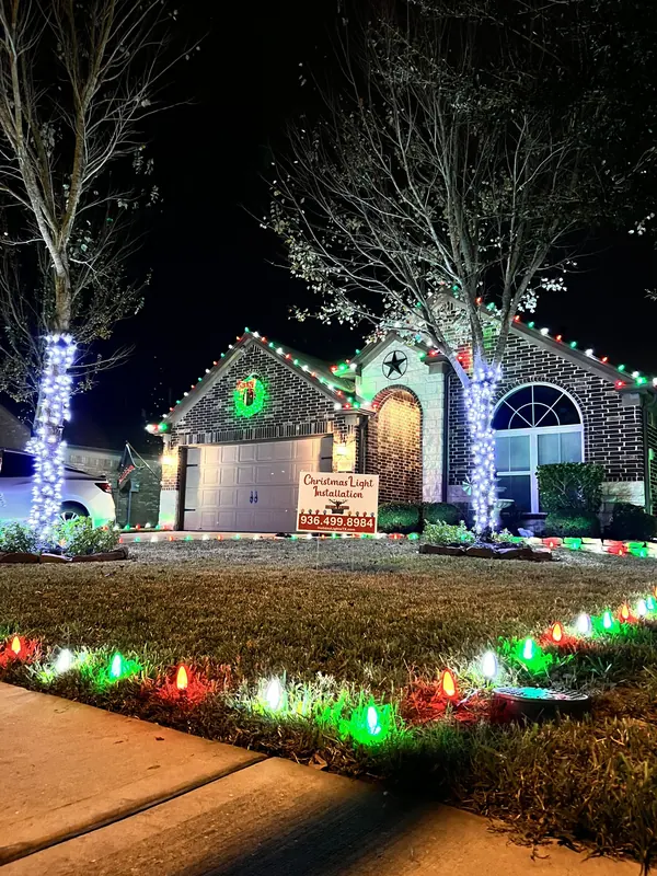 Brick ranch home at night with red, green and white roofline lights, wrapped tree trunks, a lit wreath over the garage and C9 ground lights in the lawn.