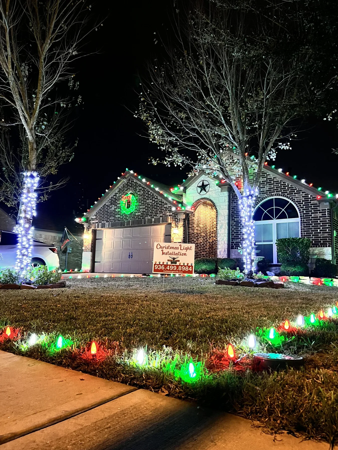 Brick ranch home at night with red, green and white roofline lights, wrapped tree trunks, a lit wreath over the garage and C9 ground lights in the lawn. Representative of installs like those near Splendora.