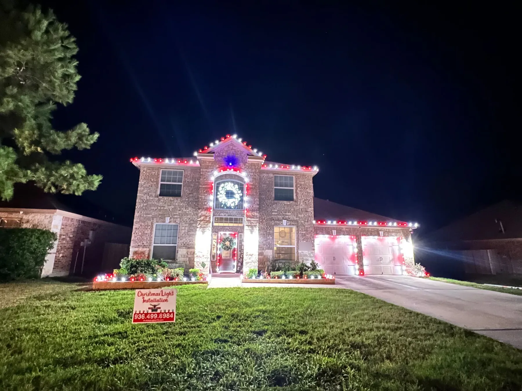 Two-story brick home with alternating red and white lights on the rooflines, a lit wreath in the arched window, outlined garage doors and lit flower beds. Representative of installs like those near Klein.