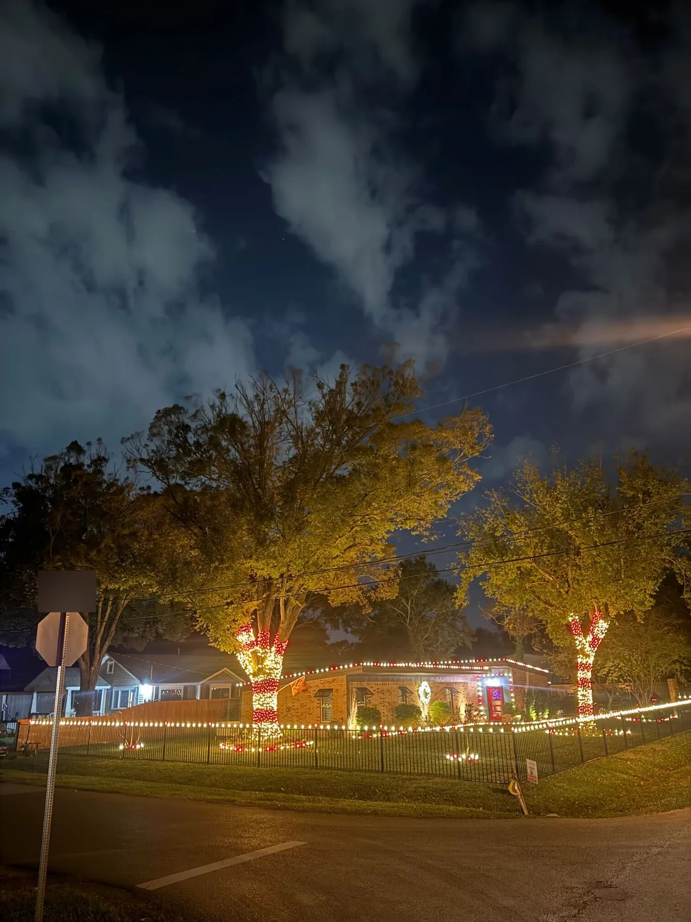 Corner-lot brick ranch at night with candy-cane red and white wrapped tree trunks, a lit roofline and a warm-white run along the iron fence. Representative of installs like those near Willis.