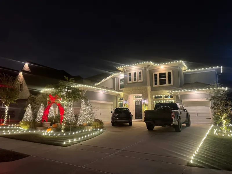 Two-story gray and limestone home at night with warm-white rooflines, wrapped trees, C9 lights edging the lawn and a giant red bow arch.