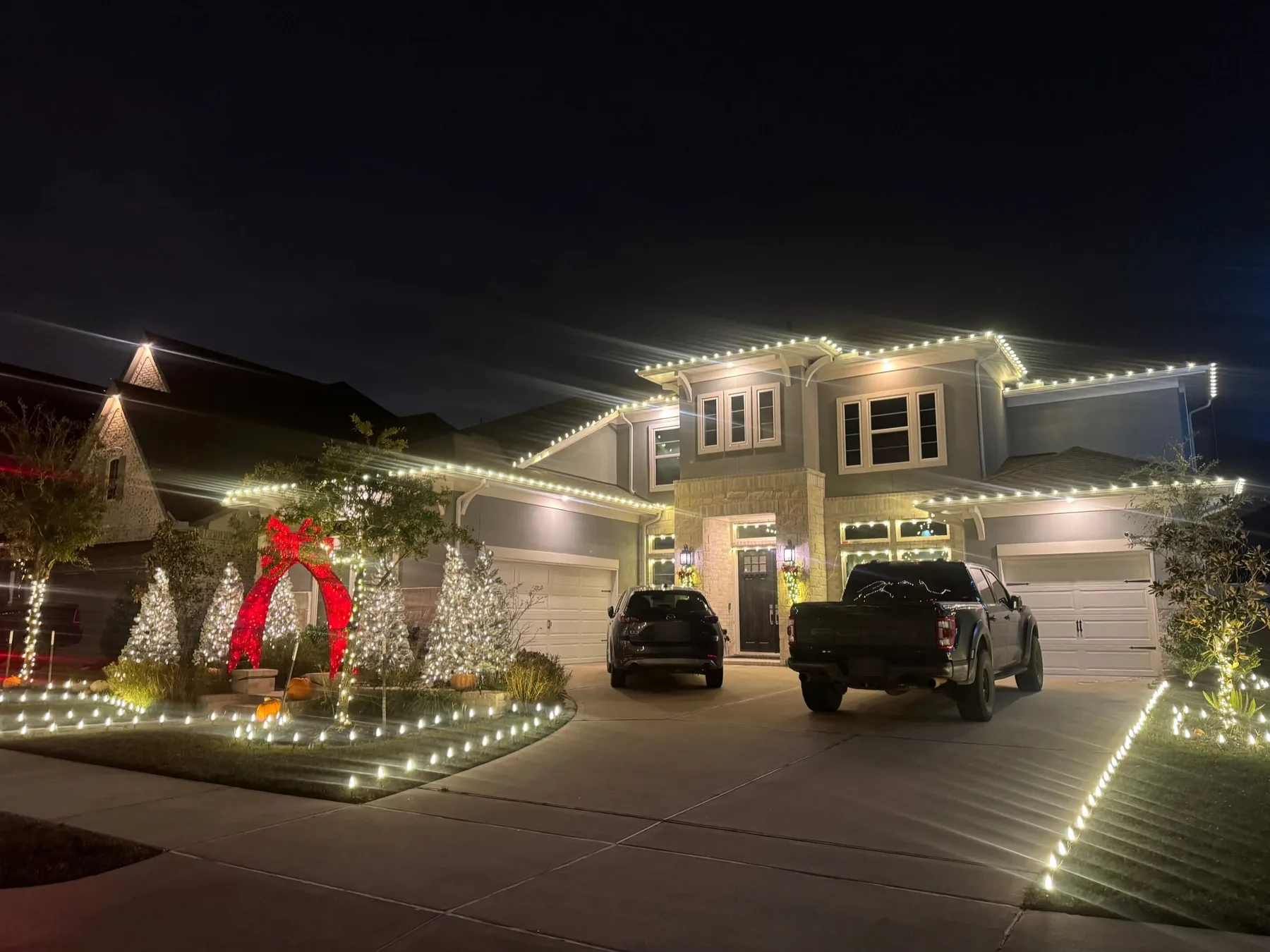 Two-story home at night with warm-white rooflines, wrapped trees and a lit lawn edge.