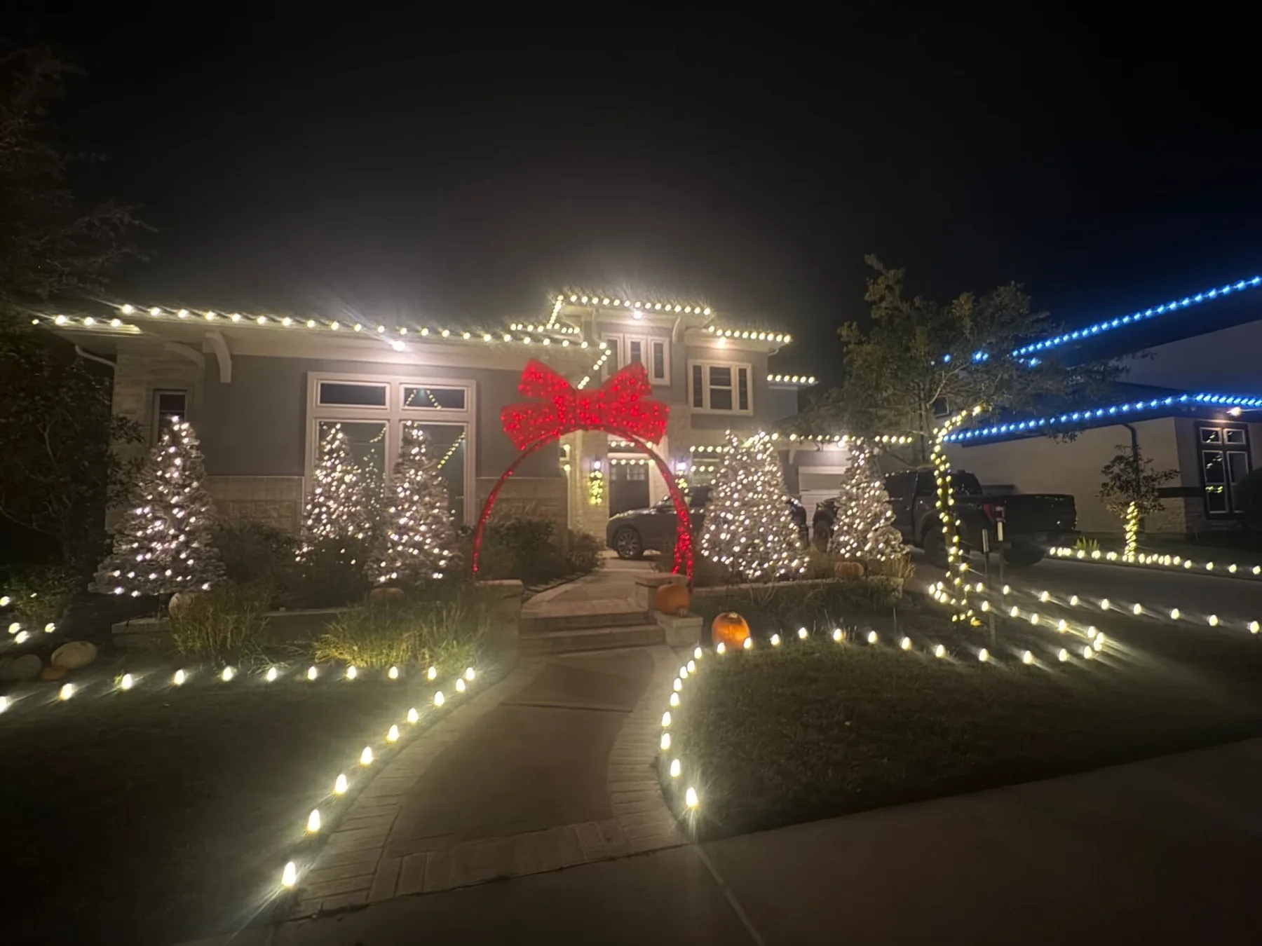 Front walkway lined with warm-white C9 ground lights leading to a red bow arch, with lit evergreens and a warm-white roofline behind. Representative of installs like those near West University Place.