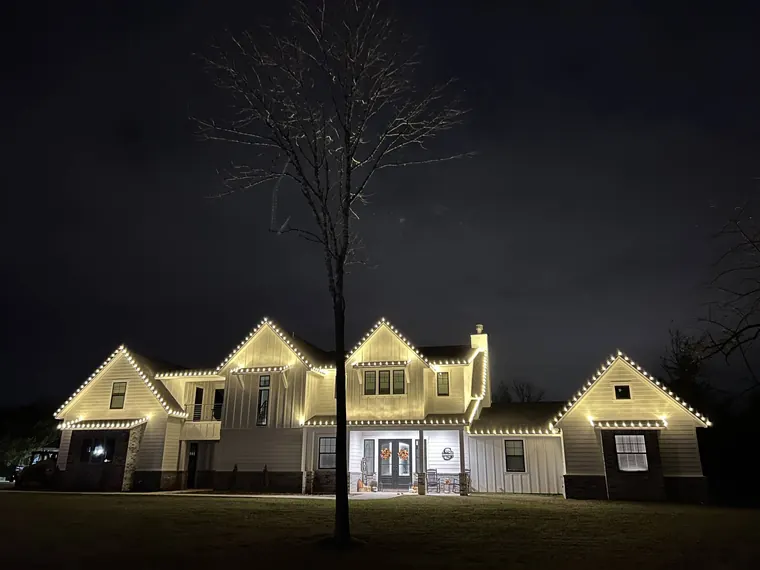 Large board-and-batten estate at night with warm-white lights tracing every gable, dormer and roofline.