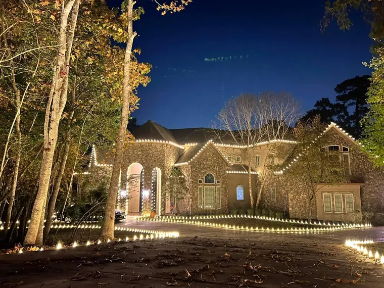 Stone estate at dusk with a lit driveway edge, wrapped tree trunks and warm-white rooflines.