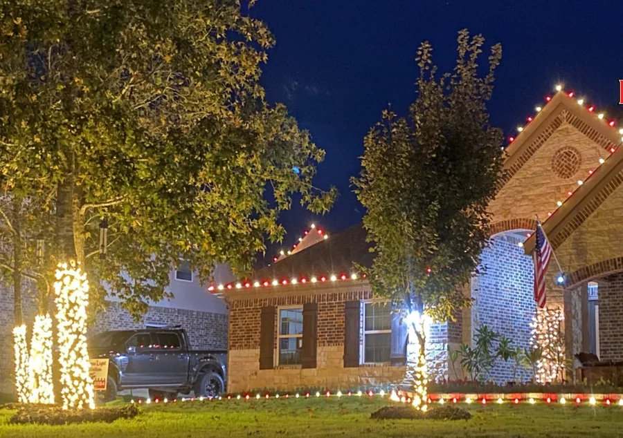 Brick home at dusk with red and white lights on the gables and warm-white wrapped tree trunks on the lawn.
