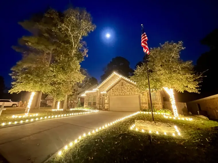 Single-story brick home under a full moon with warm-white tree wraps, a lit walkway and lit rooflines.