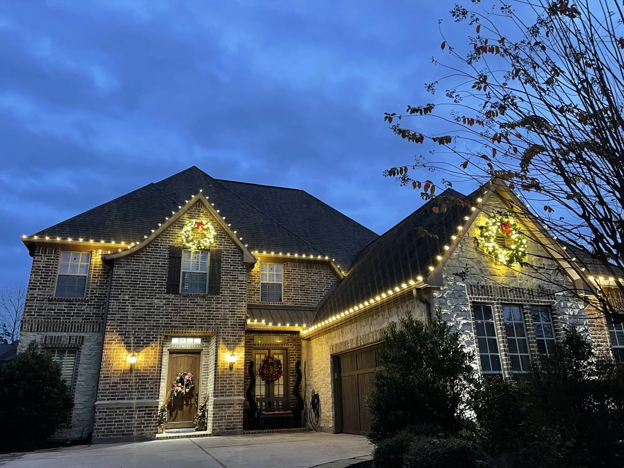 Two-storey Texas home at dusk with warm-white lights tracing the rooflines and a lit wreath on the gable.